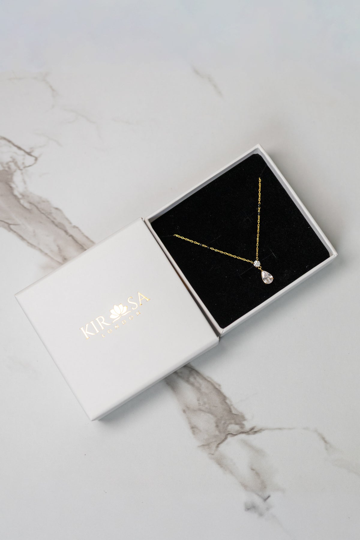 Kirosa London gold Saha Mangalsutra necklace featuring a round and pear-cut crystal drop pendant, displayed in an open white gift box on a marble surface.