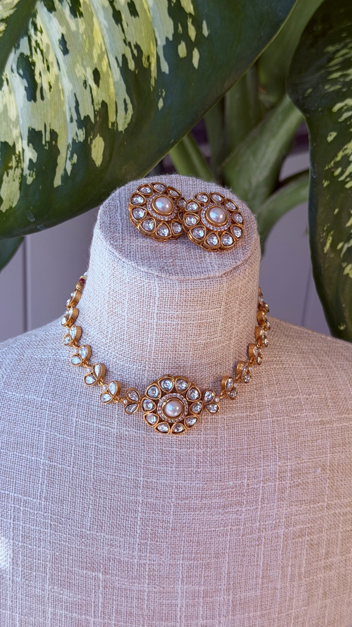 A gold-toned jewelry set displayed on a beige fabric mannequin bust, featuring a choker necklace with a central floral cluster and matching stud earrings, set against a background with green variegated leaves.