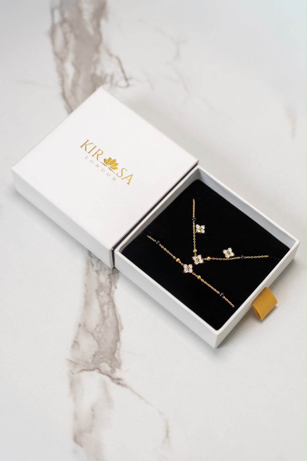 The Complete Samya Gift Set by Kirosa London featuring a gold floral crystal mangalsutra necklace, matching bracelet, and stud earrings displayed in an open white gift box on a marble surface.