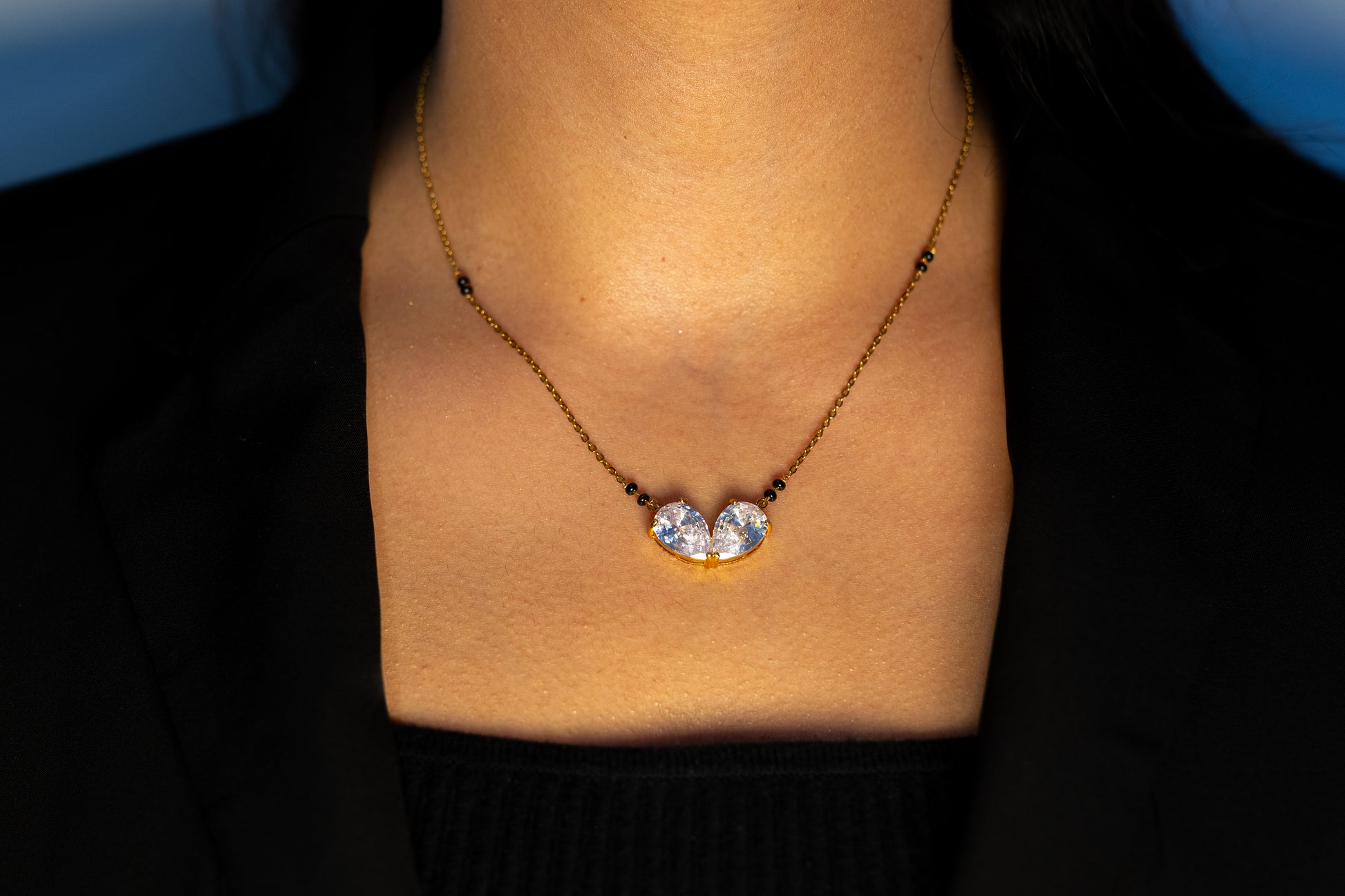 A close-up view of a woman’s neckline wearing a gold mangalsutra. The necklace has small black beads along the chain and a center pendant consisting of two large, clear teardrop-shaped stones set in gold, tilted toward each other at the base.