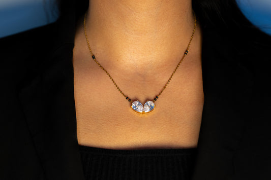 A close-up view of a woman’s neckline wearing a gold mangalsutra. The necklace has small black beads along the chain and a center pendant consisting of two large, clear teardrop-shaped stones set in gold, tilted toward each other at the base.