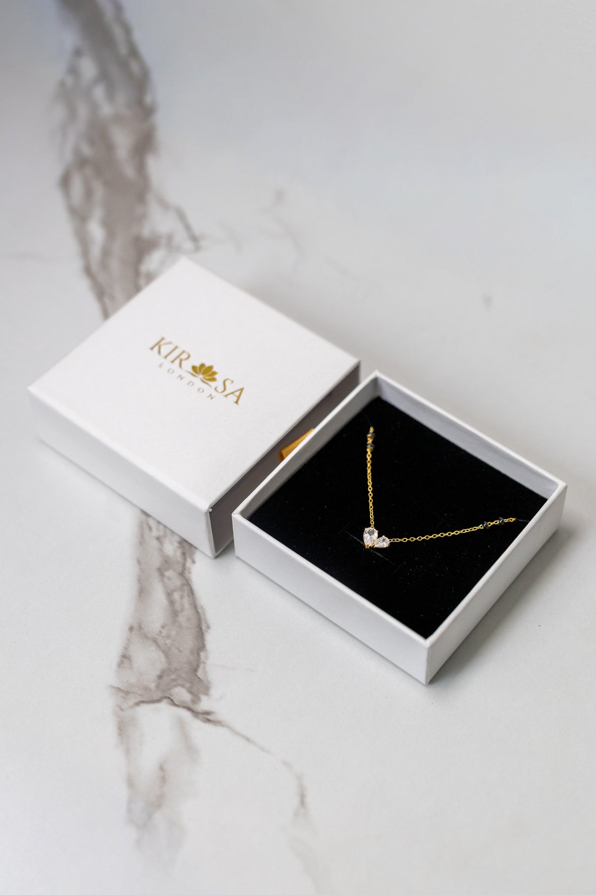 Kirosa London gold mangalsutra necklace featuring a heart-shaped dual pear-cut crystal pendant, displayed in an open white gift box on a marble surface.