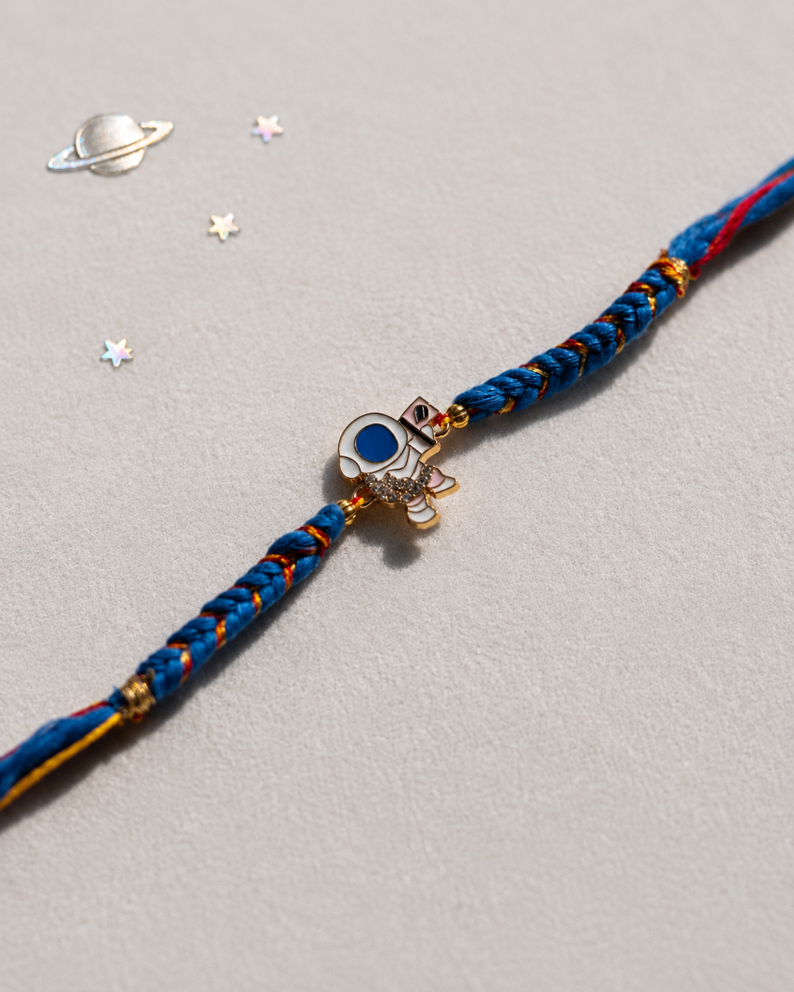 A braided blue and red cord bracelet with a gold astronaut charm featuring a blue circular detail, displayed on a textured light gray background with small decorative star and planet stickers.