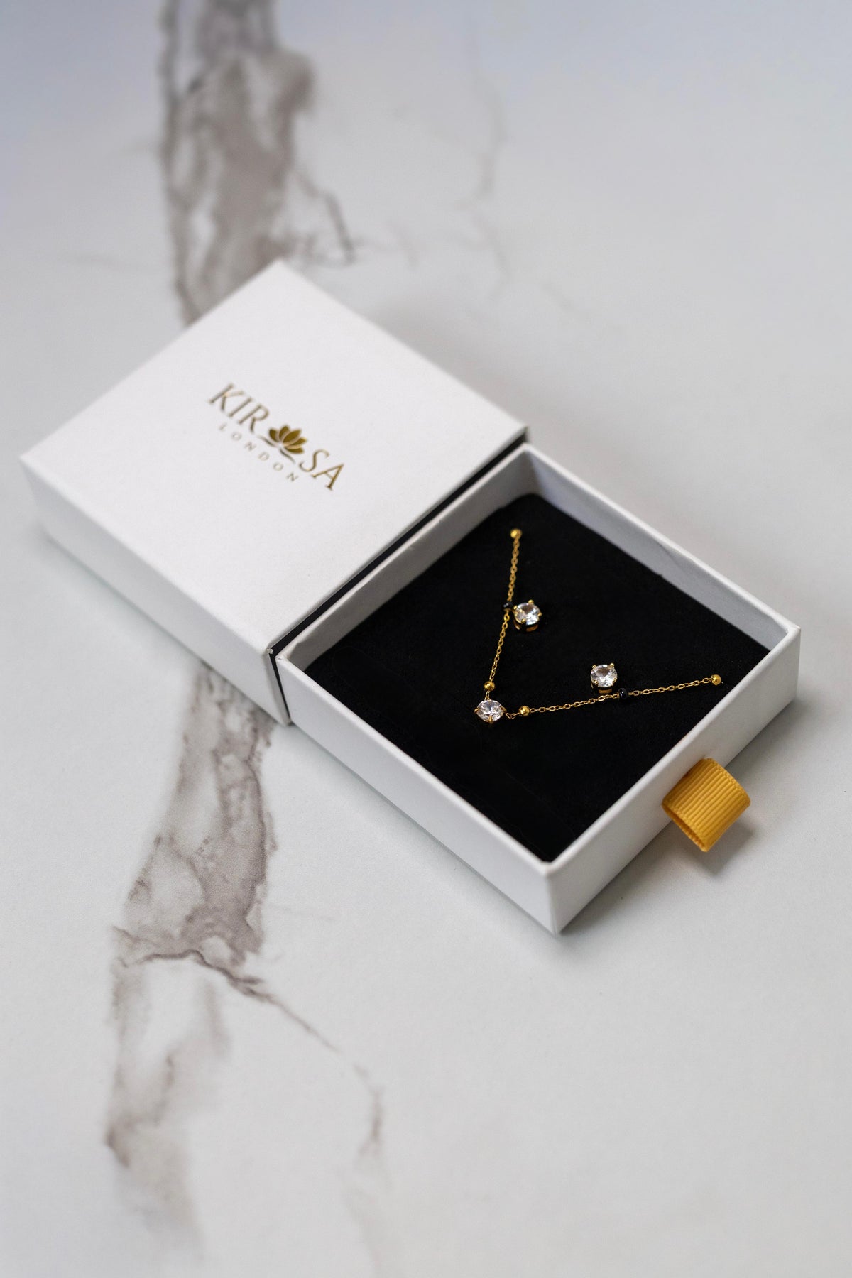The Avira Necklace and Earrings Set by Kirosa London featuring a gold solitaire mangalsutra necklace and matching stud earrings, displayed in an open white gift box on a marble surface.