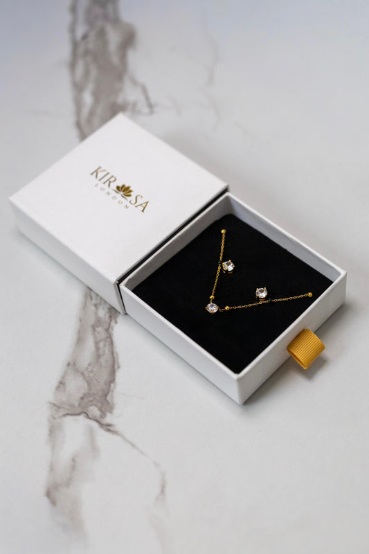 The Avira Necklace and Earrings Set by Kirosa London featuring a gold solitaire mangalsutra necklace and matching stud earrings, displayed in an open white gift box on a marble surface.