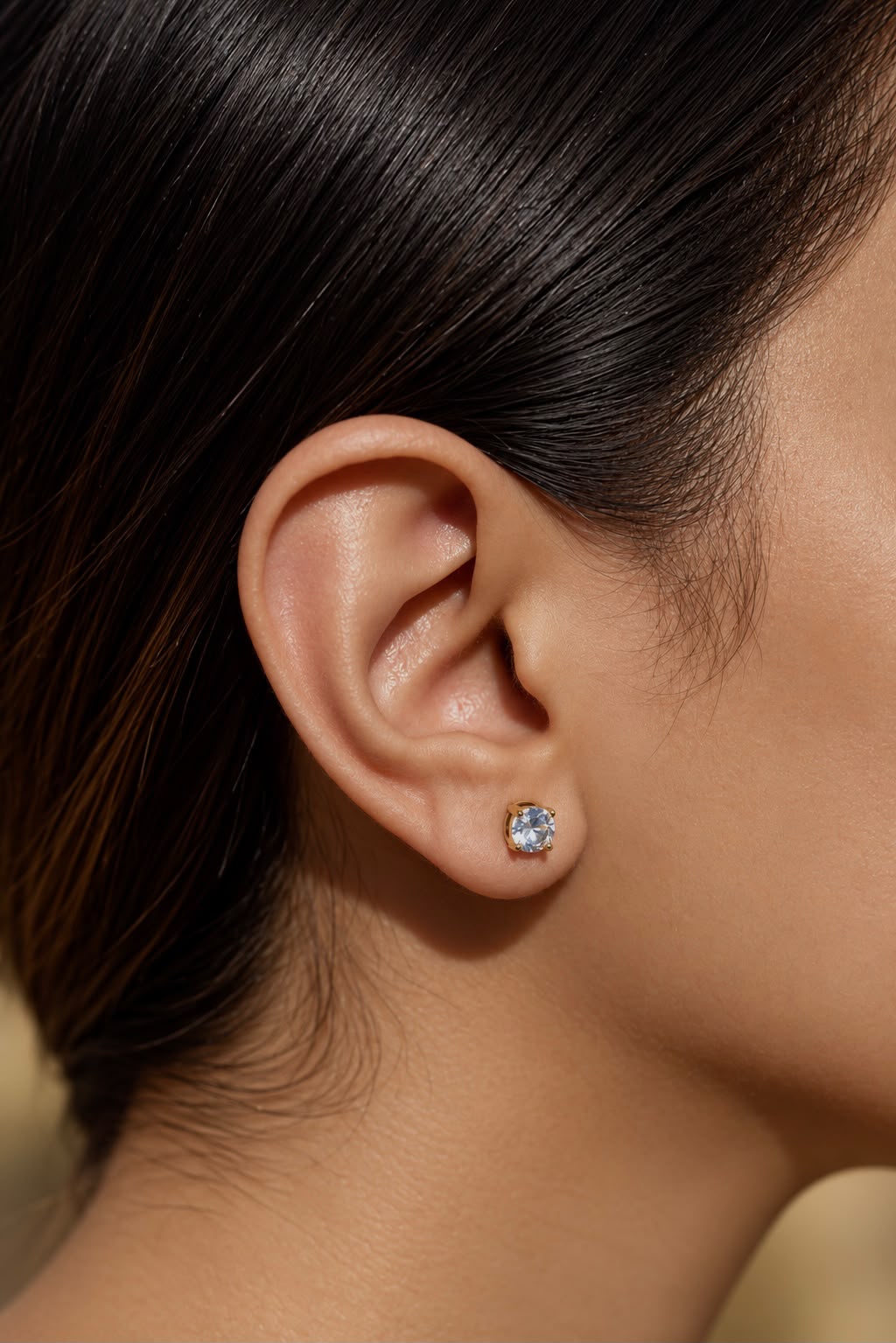 A close-up of a model's ear wearing a small round stud earring with a clear sparkling gemstone in a silver-toned setting.