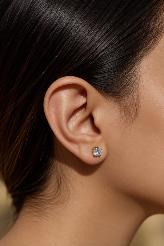 A close-up of a model's ear wearing a small round stud earring with a clear sparkling gemstone in a silver-toned setting.