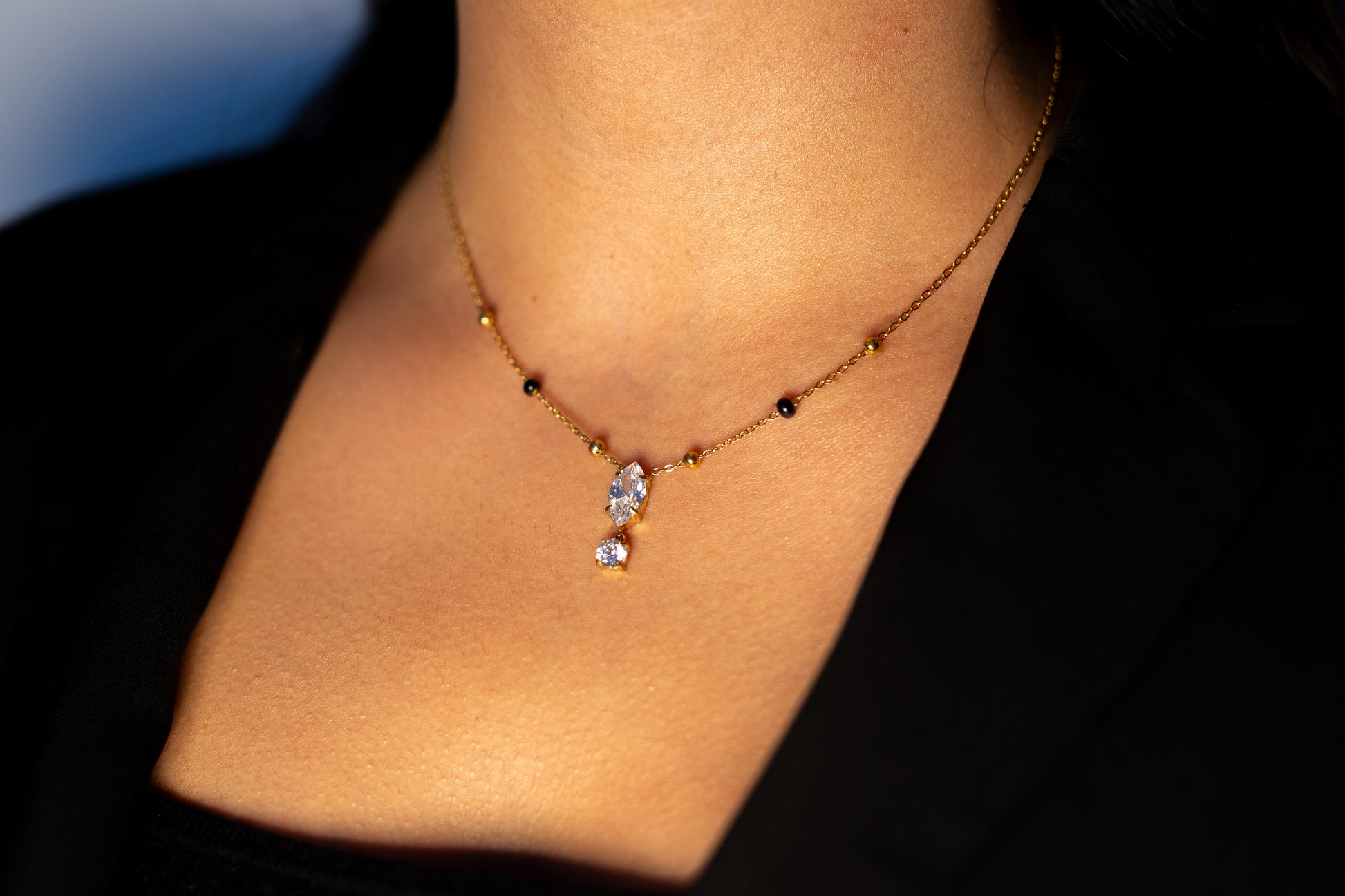 A woman wearing a black blazer and a delicate gold mangalsutra necklace. The chain has small black beads spaced along it, leading down to a pendant with a marquise-cut teardrop stone stacked above a round clear stone.
