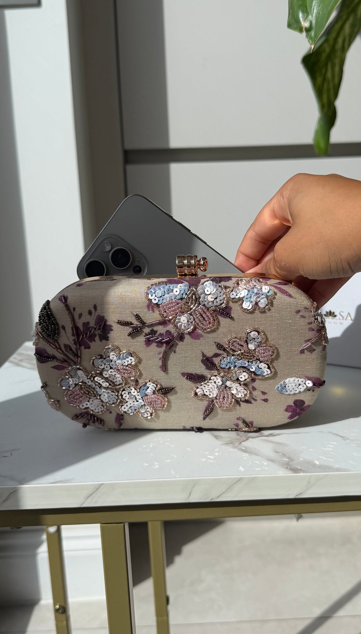 A hand holding a beige clutch bag decorated with purple and silver sequined floral patterns, with a smartphone tucked inside, set on a white marble table.