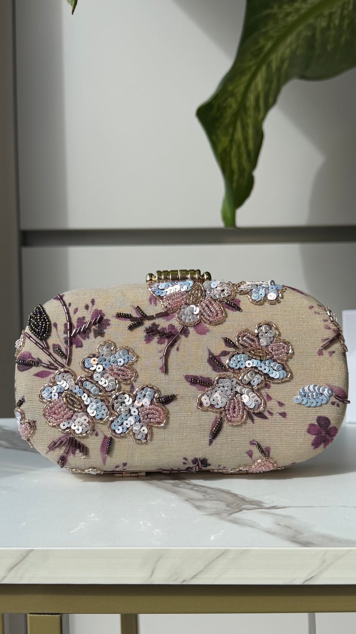 A beige clutch purse with intricate floral embroidery and sequined embellishments in pink, purple, and silver, resting on a white marble surface.