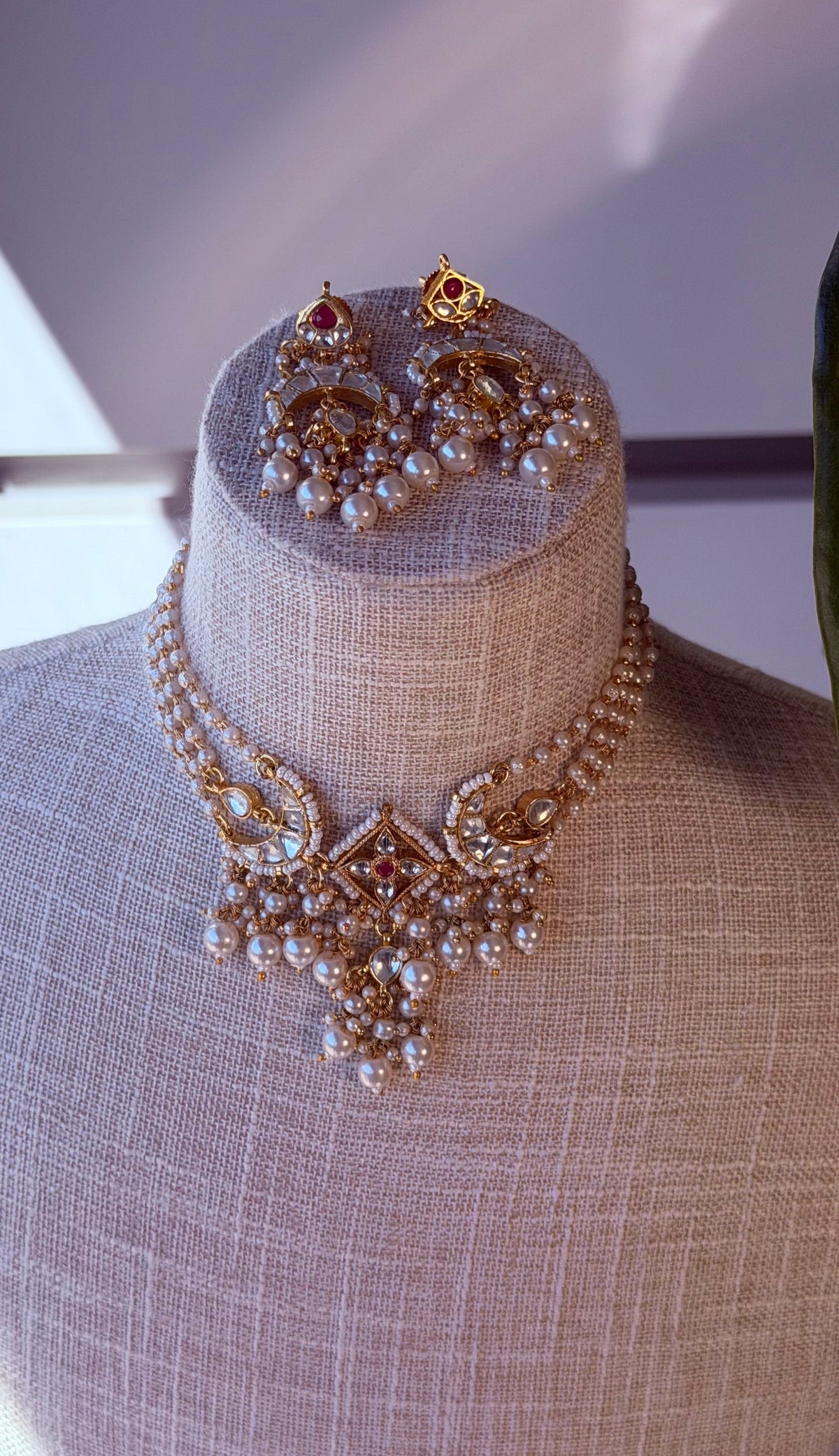 Gold and pearl traditional Indian jewelry set including a statement necklace, matching earrings, and a small stackable necklace, displayed on a beige fabric mannequin bust.