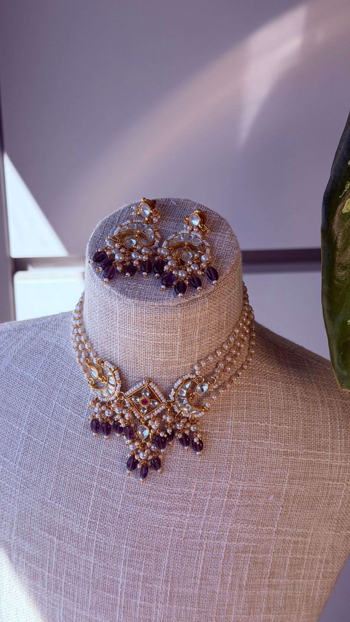 An ornate gold-toned traditional necklace and matching earrings displayed on a beige fabric mannequin bust. The jewelry features intricate beadwork, clear stones, and dark purple dangling accents.