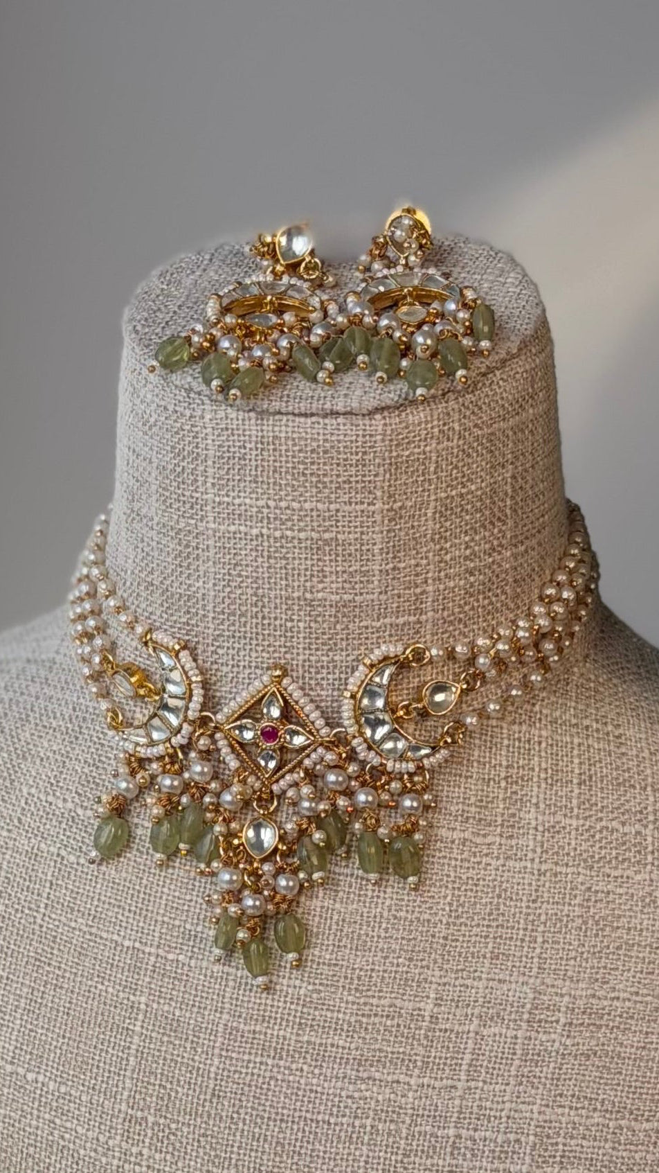 Gold and light green (mint green) beaded kundan jewelry set displayed on a beige linen jewelry bust, featuring a statement necklace and matching earrings, set against a neutral background with a green plant leaf on the side.