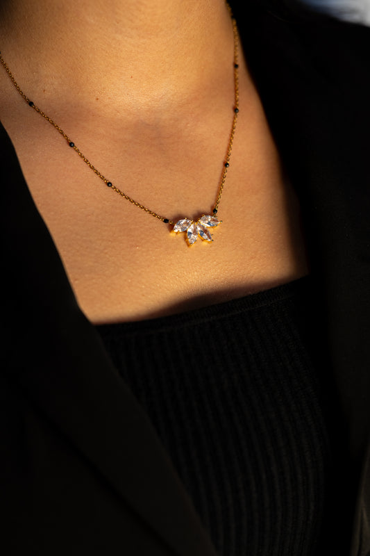A woman wearing a black top and blazer, beautifully illuminated by warm light, showcasing a delicate gold mangalsutra necklace. The chain has small black beads and features a center pendant designed like a blooming lotus flower with five clear, pointed stones.