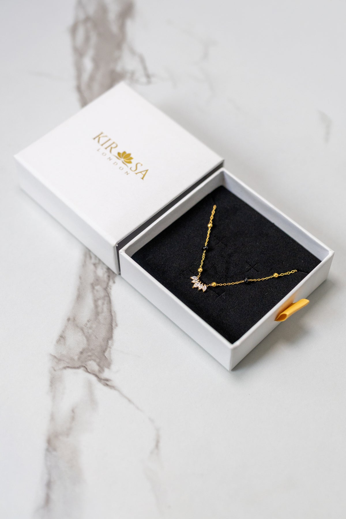 Kirosa London gold Padma 2.0 mangalsutra necklace featuring a lotus-inspired marquise crystal cluster pendant, displayed in an open white gift box on a marble surface.