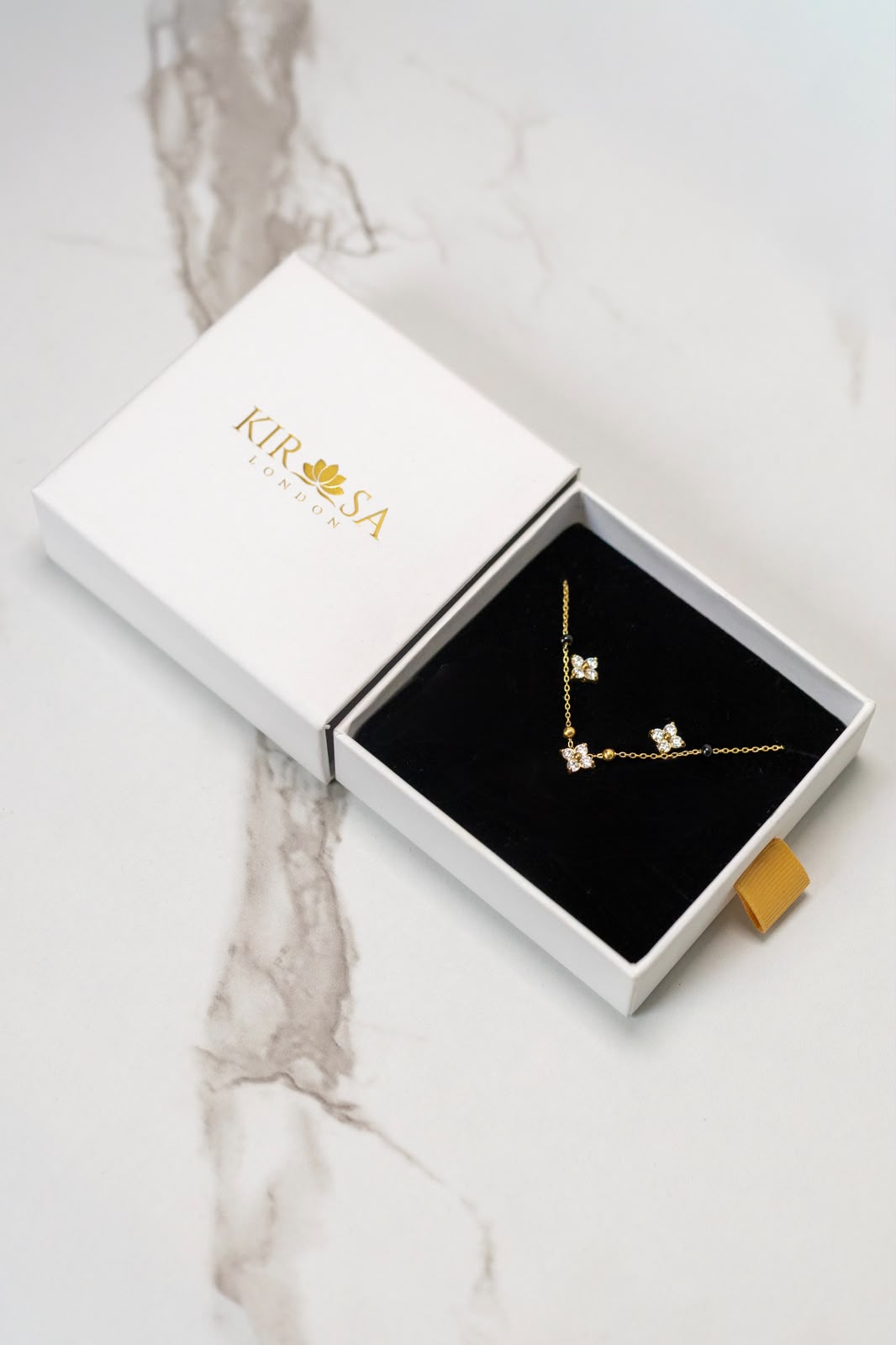 The Samya Necklace and Earrings Set by Kirosa London featuring a gold floral crystal mangalsutra necklace and matching stud earrings displayed in an open white gift box on a marble surface.
