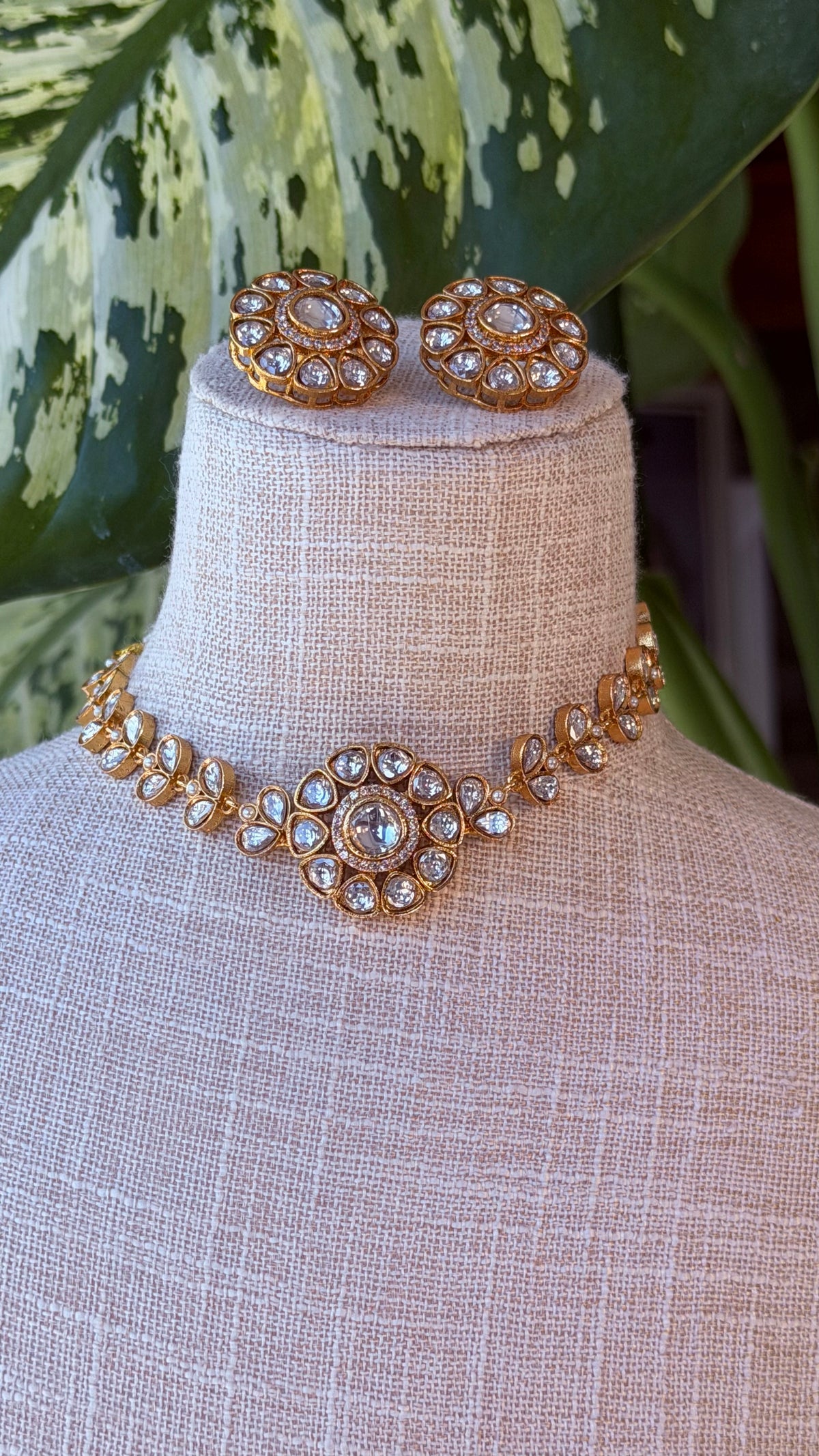 Gold-toned jewelry set displayed on a beige fabric mannequin neck, featuring a choker necklace and matching stud earrings with clear rhinestone-like stones, set against a leafy background.