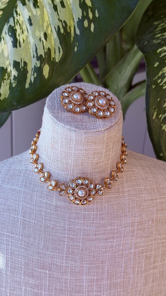 A gold-toned jewelry set displayed on a beige fabric mannequin bust, featuring a choker necklace with a central floral cluster and matching stud earrings, set against a background with green variegated leaves.