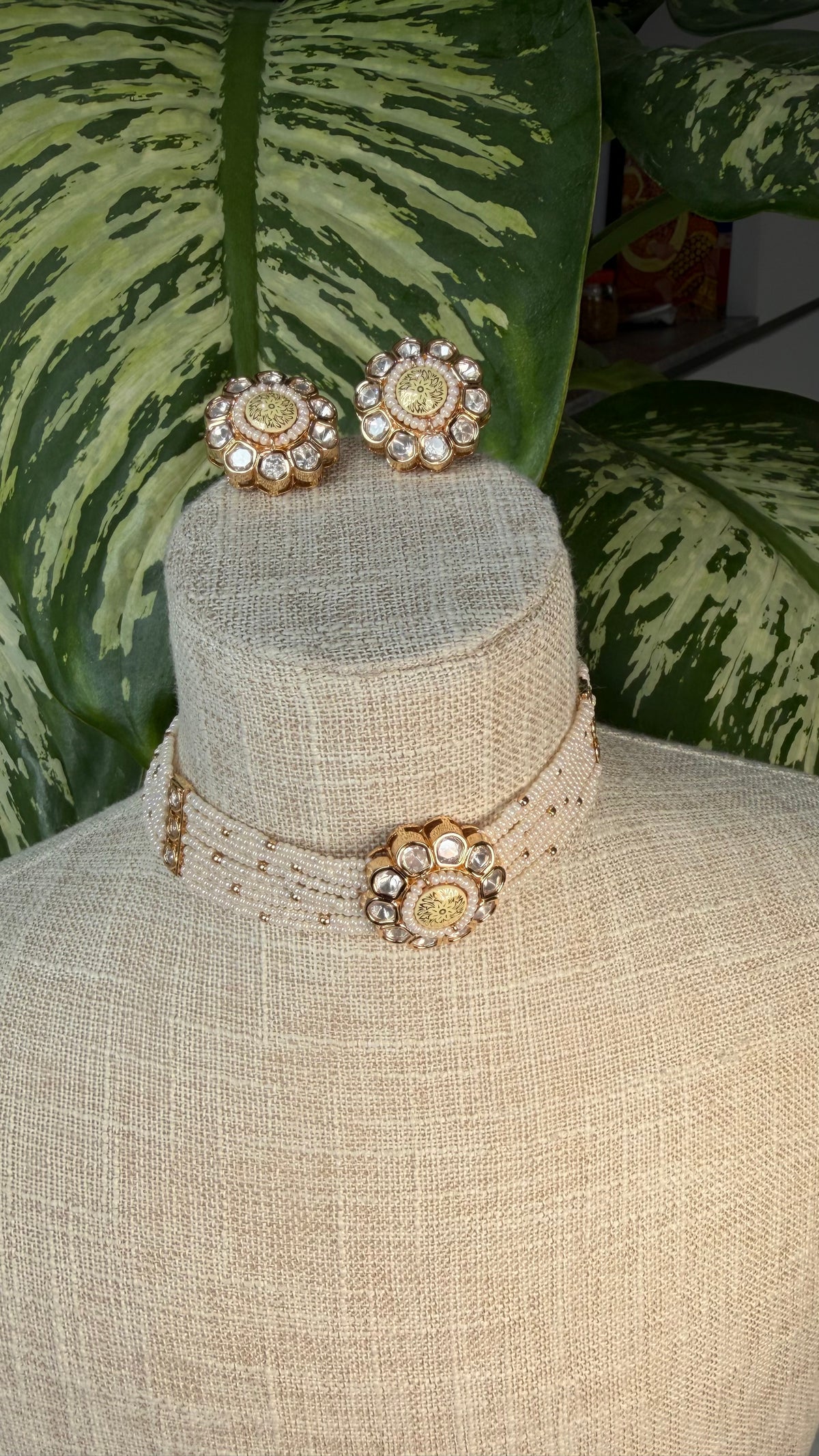 A gold-toned kundan jewelry set displayed on a beige fabric mannequin bust, featuring a beaded choker necklace with a central floral embellishment and matching stud earrings, set against a background of green variegated houseplant leaves.