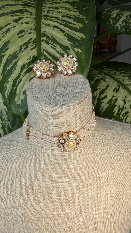 A gold-toned kundan jewelry set displayed on a beige fabric mannequin bust, featuring a beaded choker necklace with a central floral embellishment and matching stud earrings, set against a background of green variegated houseplant leaves.
